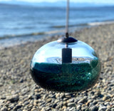 A blue-green recycled glass pendant in the background of a rocky beach.