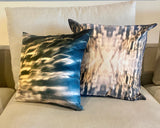 Two square-shaped silk throw pillows with a geometric pattern featuring blue and orange light effects, displayed on chair