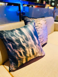 Two square-shaped silk throw pillows with a geometric pattern featuring blue and orange light effects, displayed couch to the side