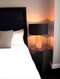 A table lamp with a rose quartz glass base and a charcoal-colored shade with a copper interior, complemented by a black linen shade in a bedroom scene