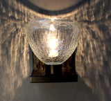 A ACQUA clear sconce with crackled glass in a dark room, creating a speckled light effect on the white wall it is mounted to.