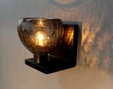 A close-up side angle of a small wall light on a black metal stand made out of grey recycled glass with a cracked pattern 
