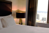 A grey glass lamp in a bedroom scene with a window looking out to the city and white bed sheets